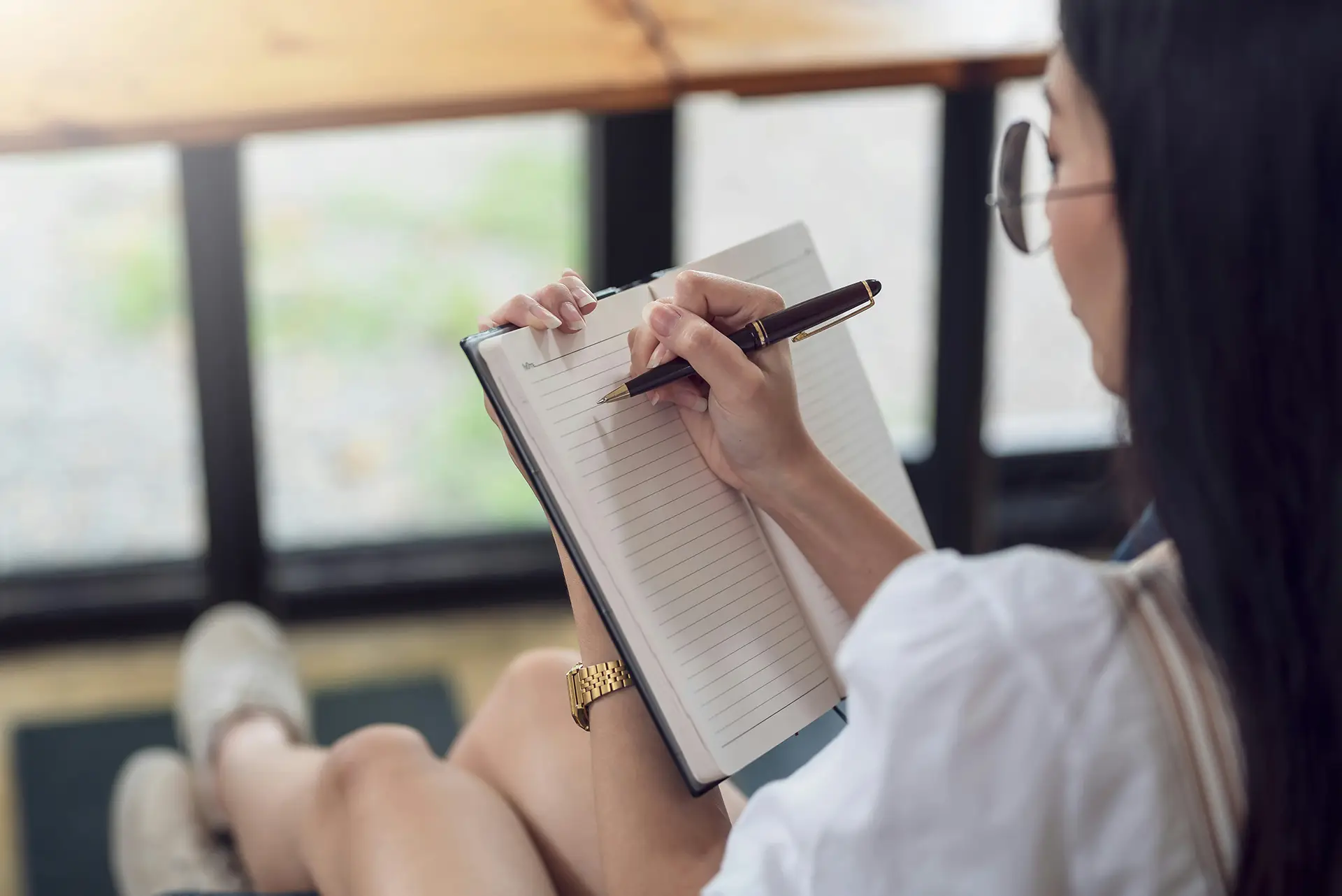 Woman with glasses writing in a blank lined notebook while sitting near a window.