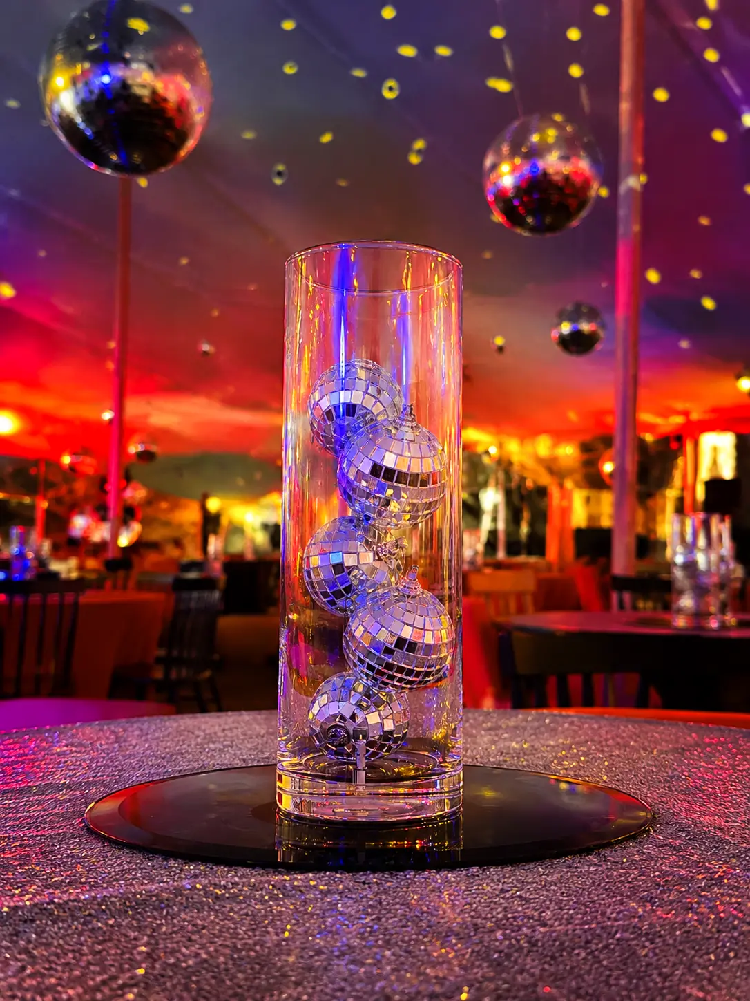 Tall clear vase with small disco balls inside on a sparkling table at a colorful party venue.