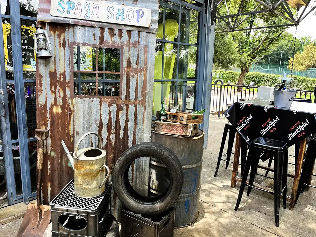 Rusty metal shop front with a watering can, tire, and crates near outdoor bar seating under trees.