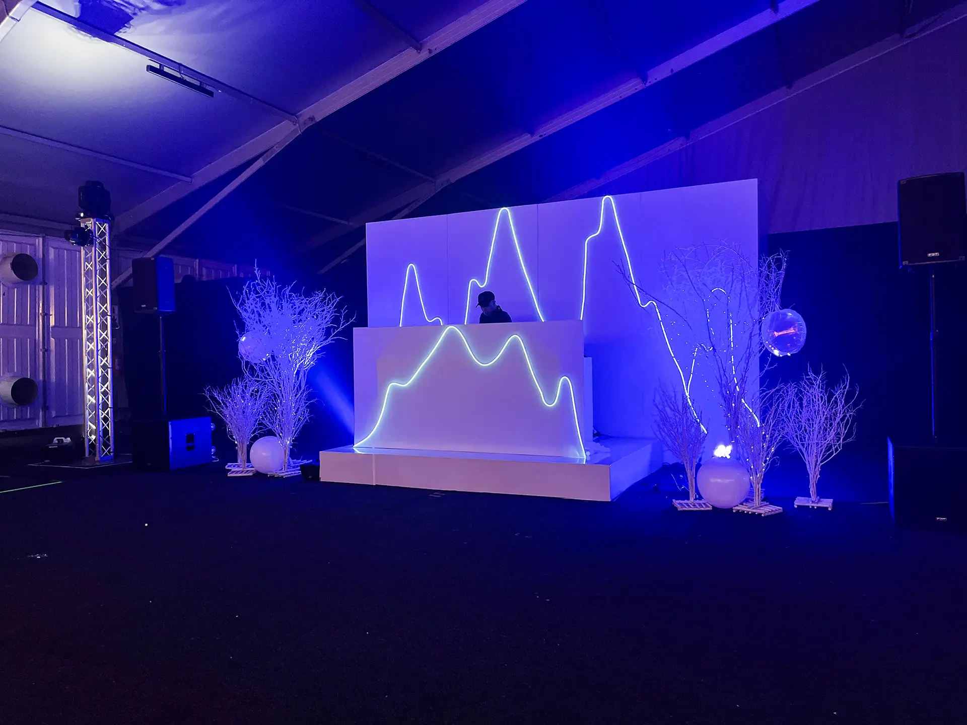 DJ booth with neon mountain light design surrounded by white decorative branches under blue lighting.