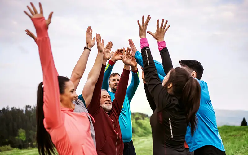 Group of six people outdoors raising their hands together in a team cheer or stretch.