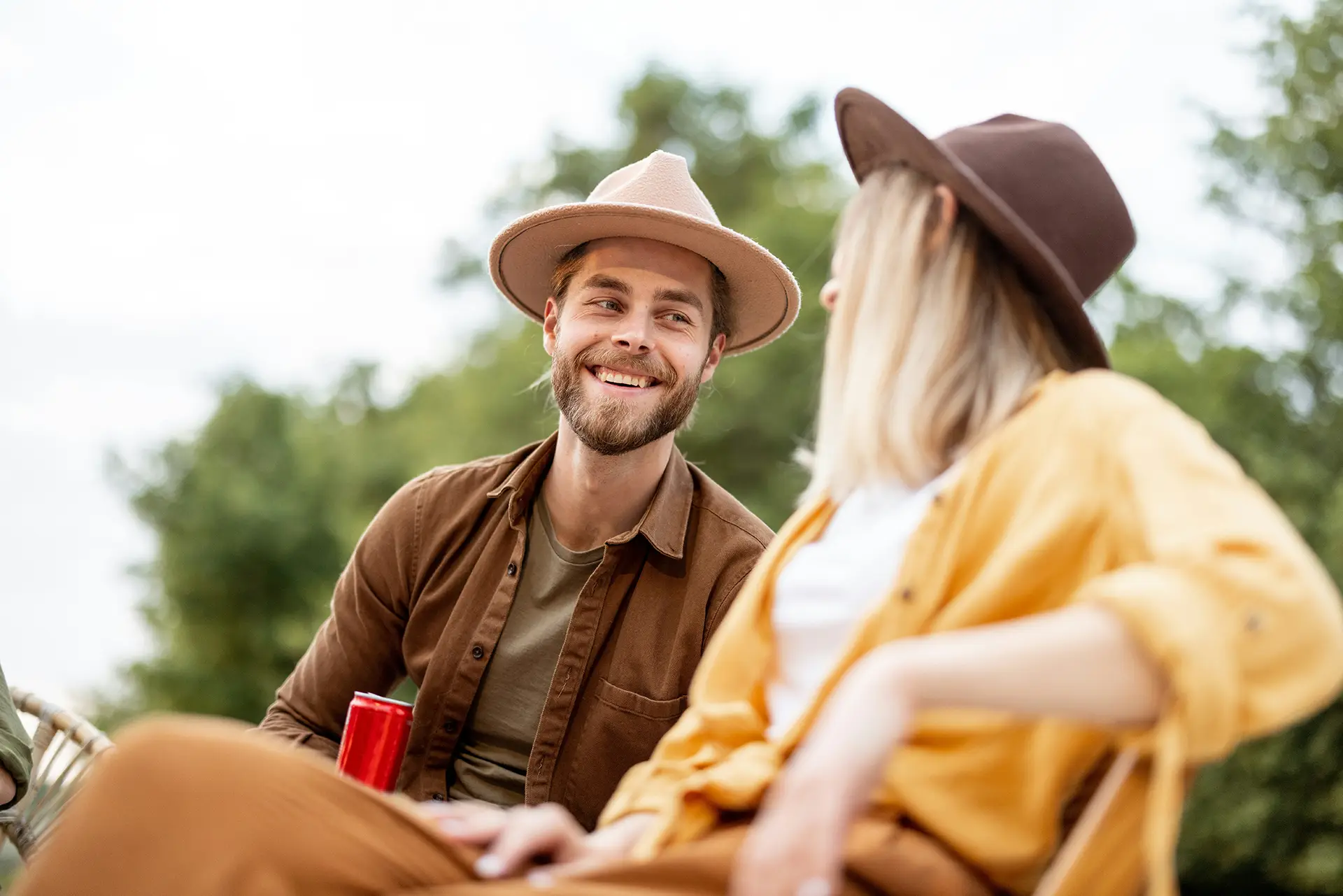 Smiling man in a hat holding a red can, talking outdoors with a woman in a yellow shirt and hat.