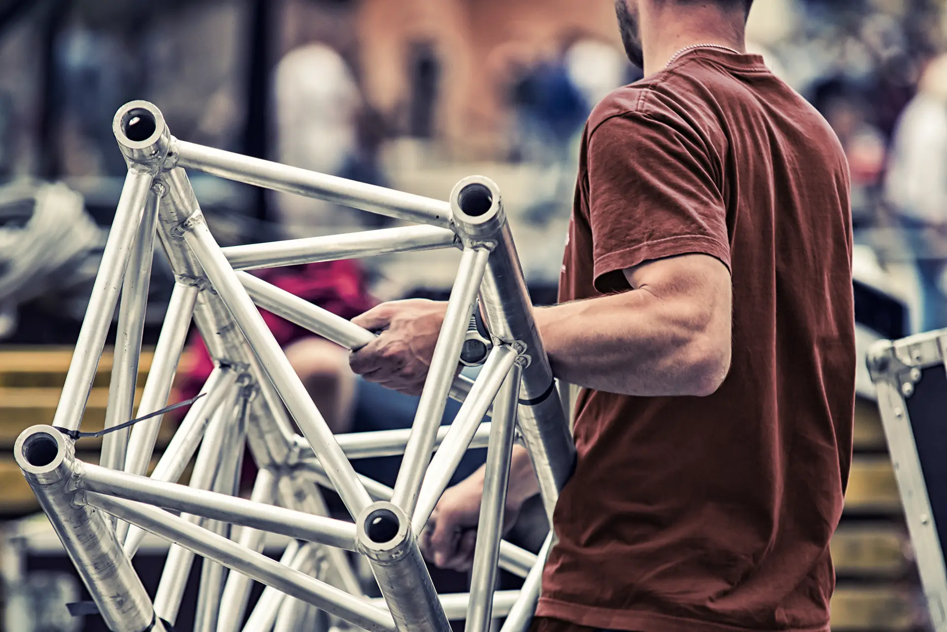 Man in a brown shirt holds a silver metal truss structure outdoors with blurred background.